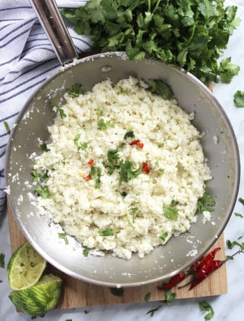 Cauliflower rice in a metal skillet with fresh cilantro, squeezed limes and red chili peppers.