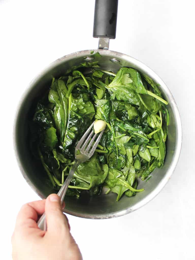 A clove of garlic on a fork stirring the spinach in a pot.