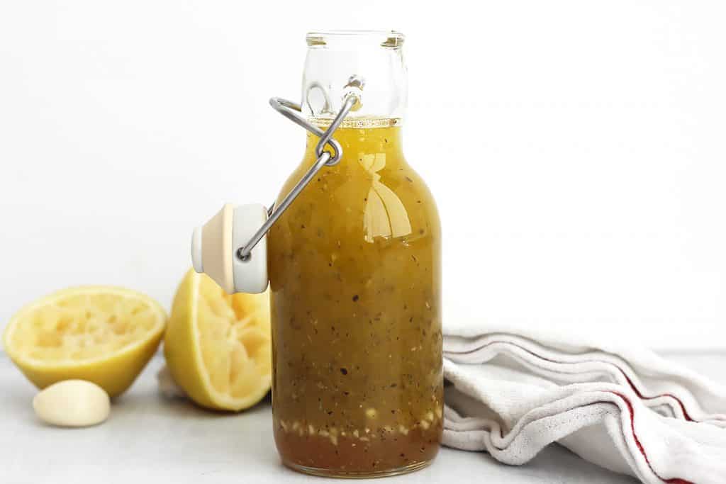 A glass bottle of honey lemon dressing next to a garlic clove and lemon halves.