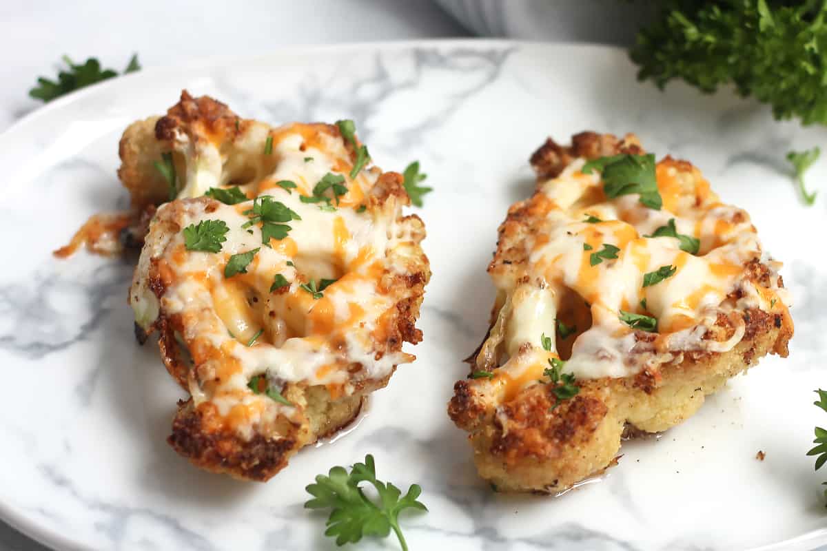 Two cheesy cauliflower steaks on a plate with fresh parsley.
