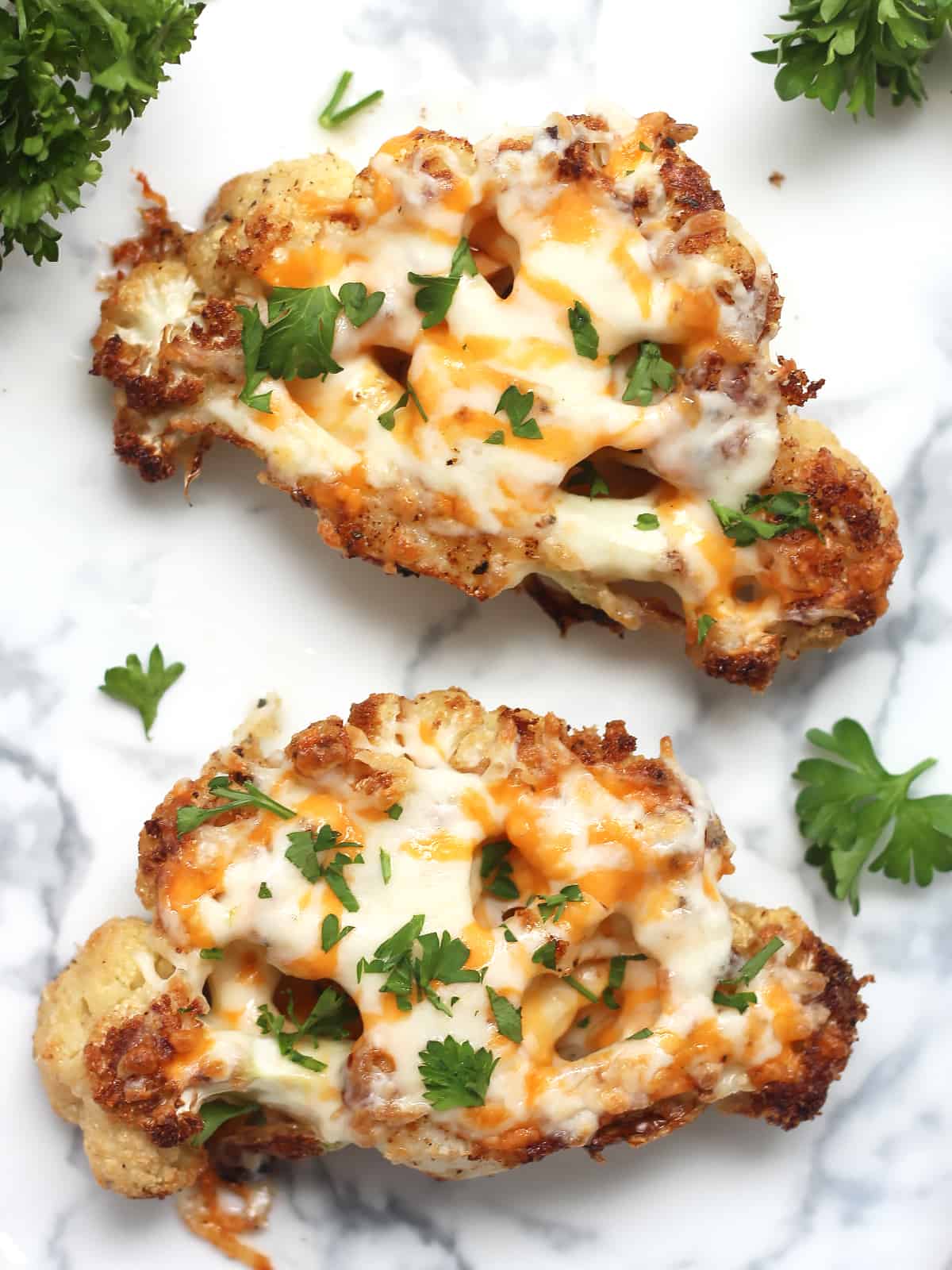 Overhead shot of two cheesy cauliflower steaks on a plate with fresh parsley.