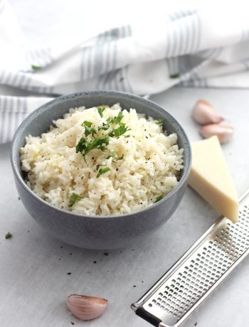 A bowl of rice next to a wedge of parmesan and garlic cloves.