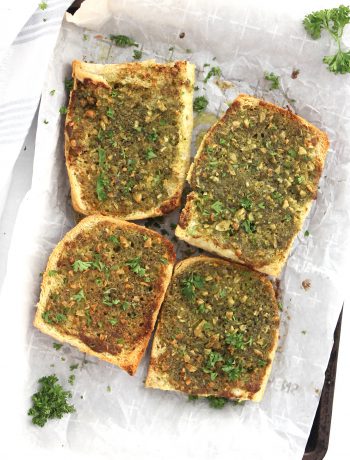 Four pieces of pesto garlic bread on a lined baking sheet with fresh parsley garnish.