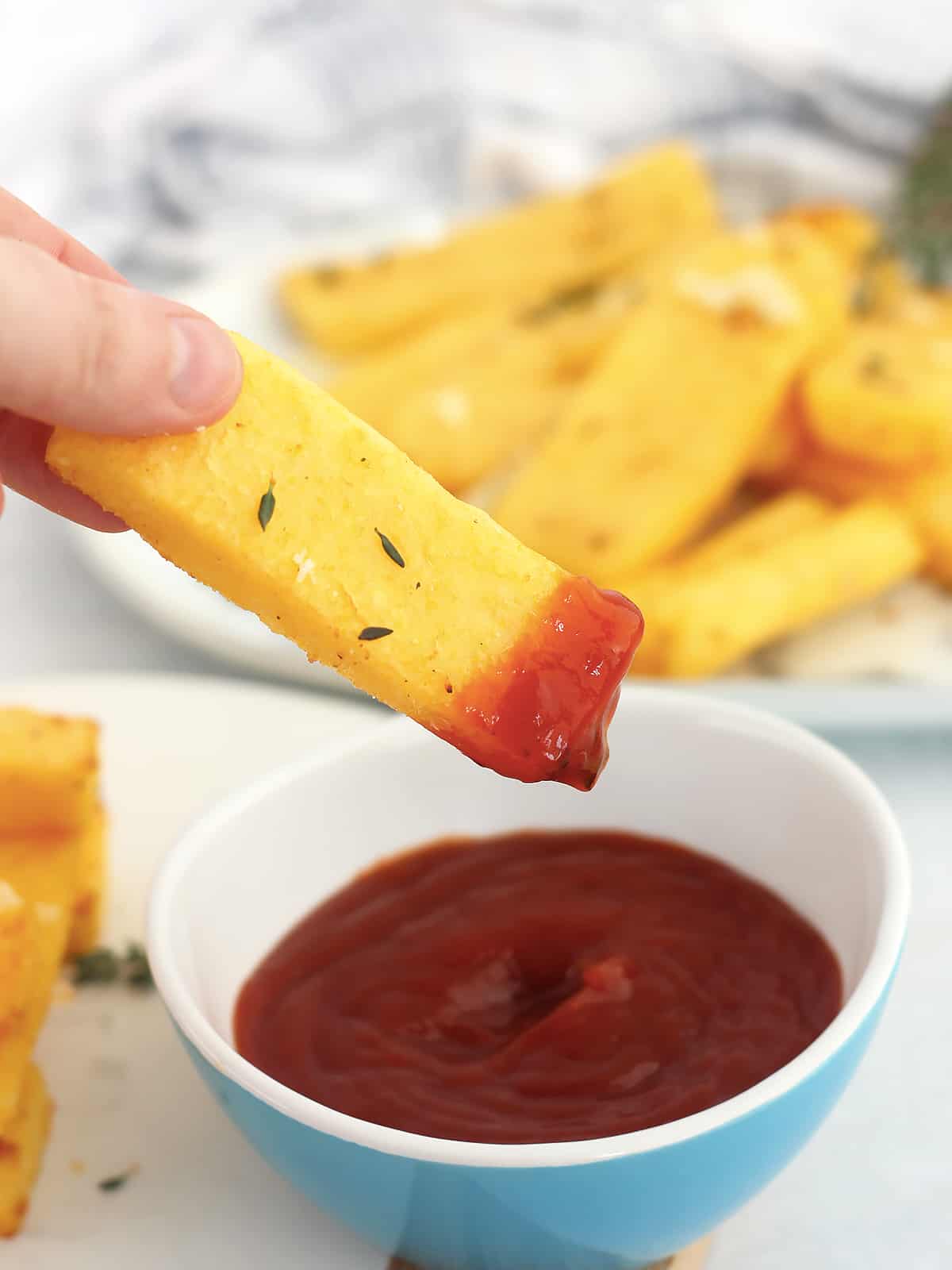 A polenta fry being dipped in a bowl of tomato ketchup.