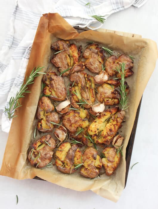 Smashed fingerlings on a lined baking sheet garnished with sprigs of rosemary.