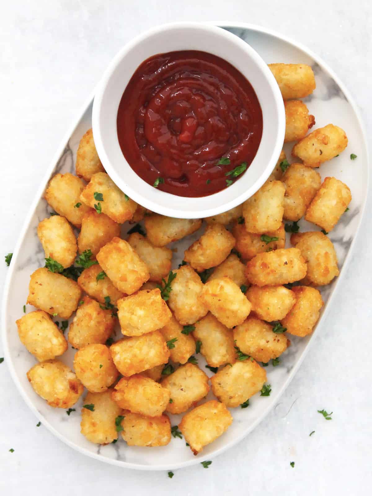 Overhead shot of air fryer tater tots on a white plate with a pot of tomato ketchup and garnished with fresh parsley.