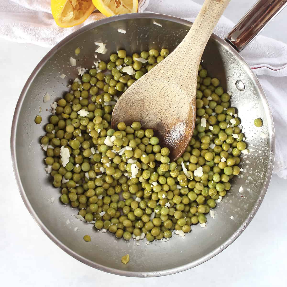 Sautéed green peas in a skillet next to two freshly squeezed lemon halves.