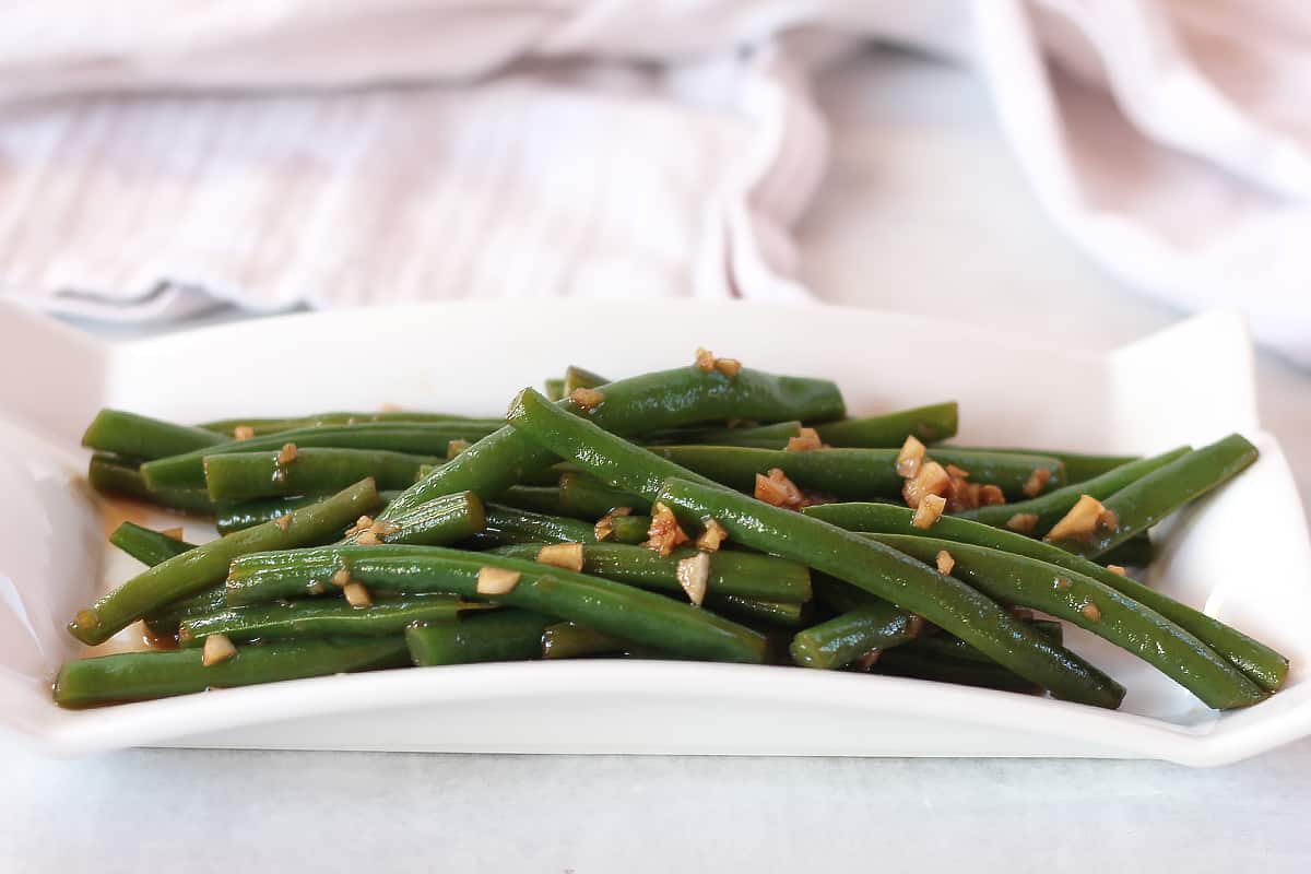 Cooked green beans served on a white plate with balsamic vinegar and garlic poured over the top.