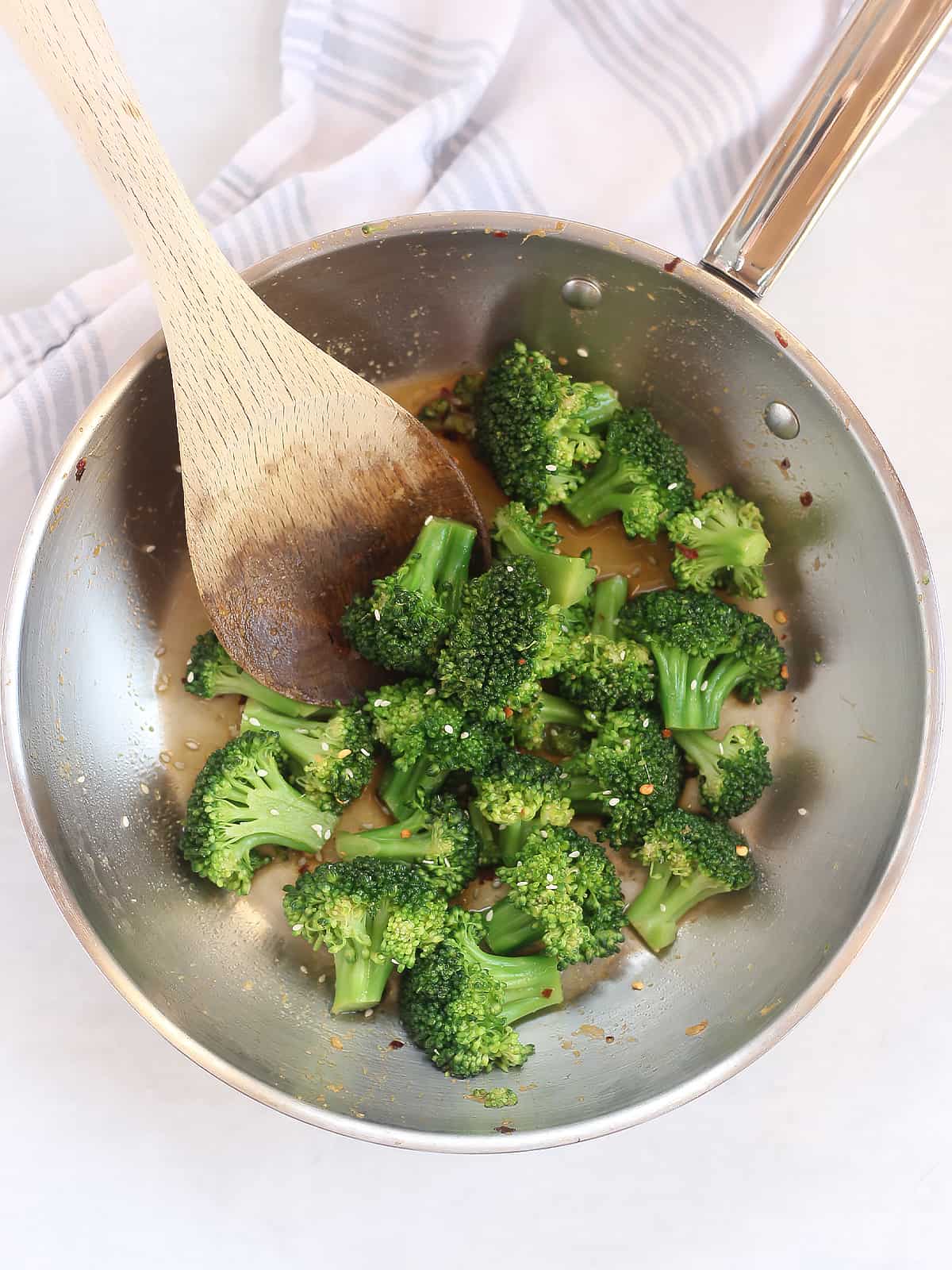 Broccoli florets in a skillet with soy sauce, being stirred with a wooden spoon.