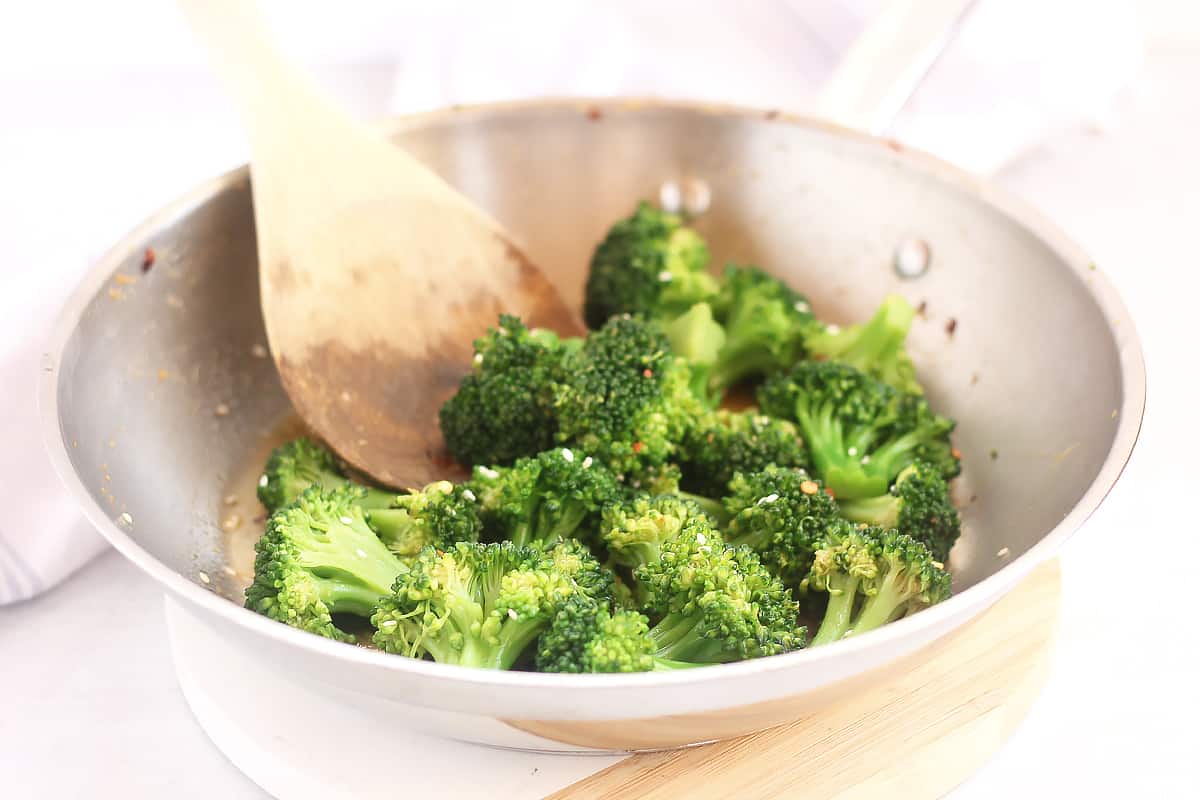A wooden spoon stirring broccoli in soy sauce in a silver skillet.