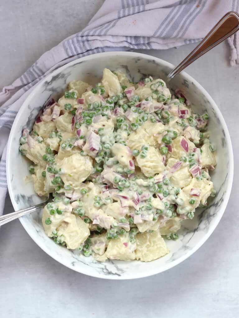 Creamy potato salad with green peas and red onions in a bowl with two spoons.