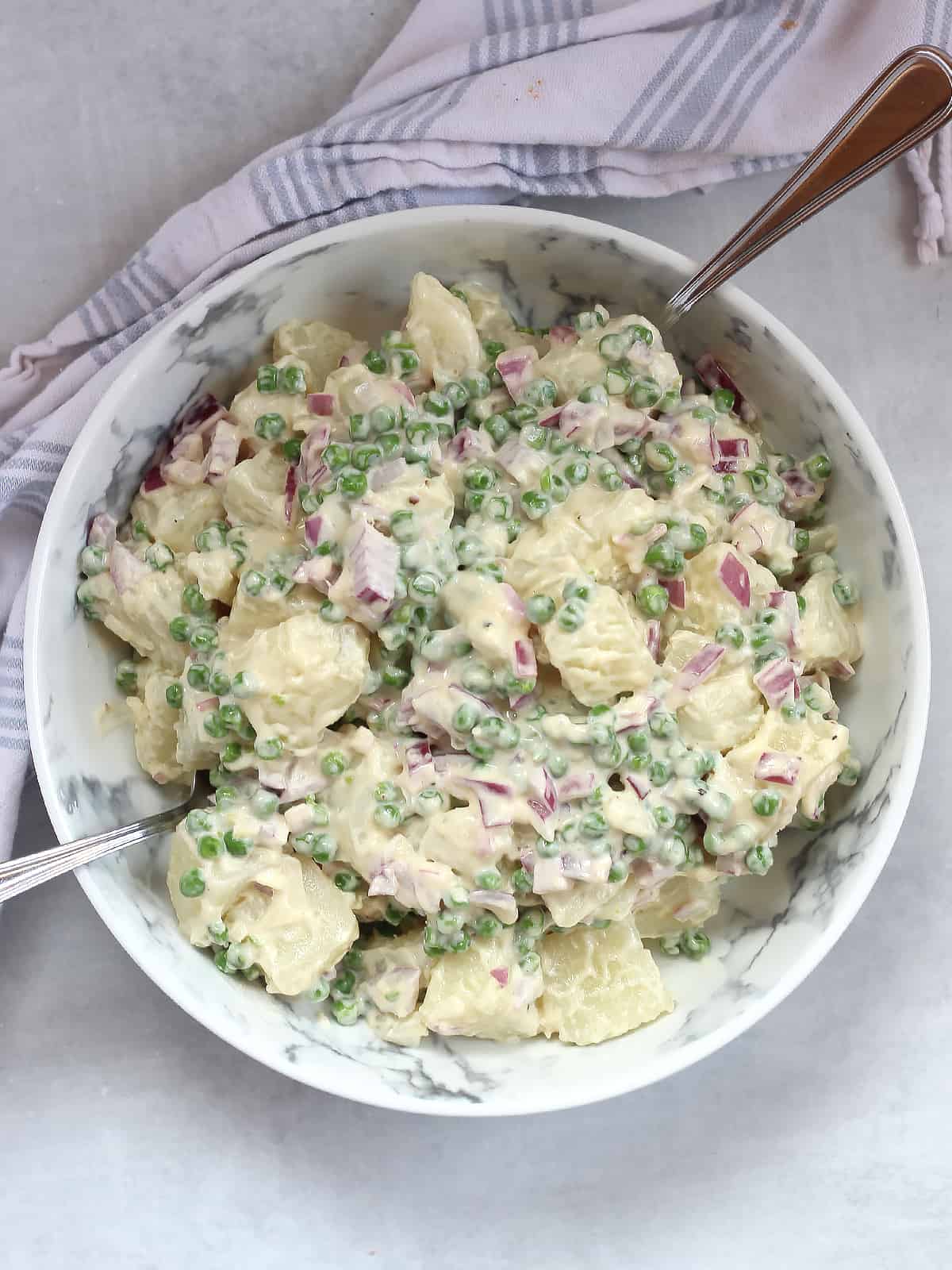 Creamy potato salad with green peas and red onions in a bowl with two spoons.