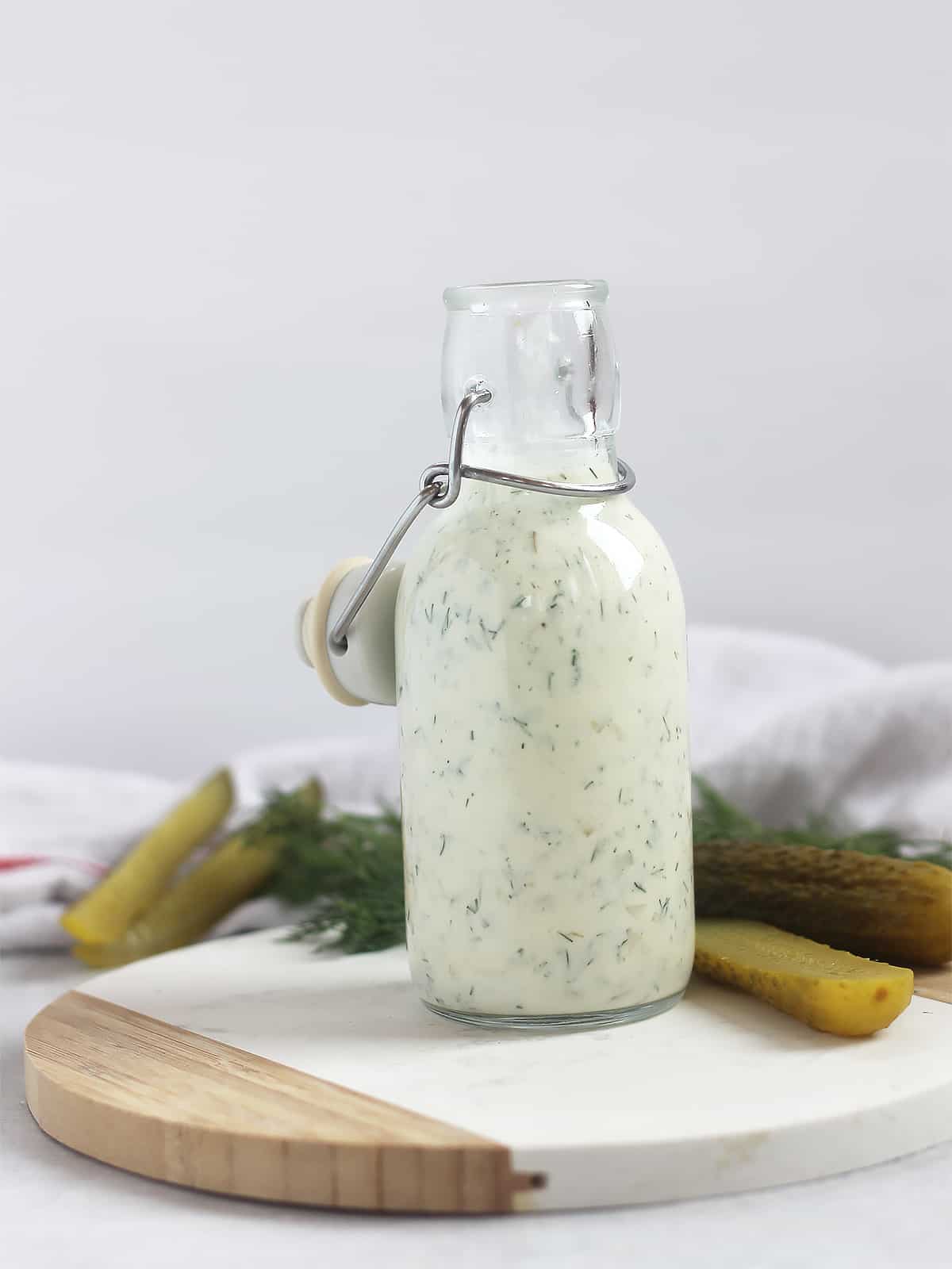 A bottle of creamy dill pickle dressing on a wooden chopping board next to fresh ingredients.