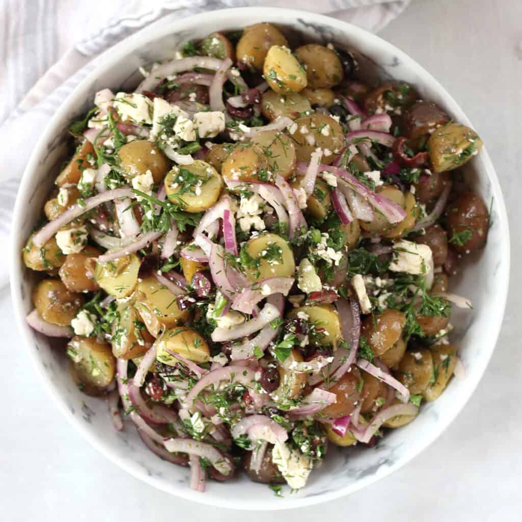 Greek potato salad with feta, olives, onions and fresh herbs in a white bowl.