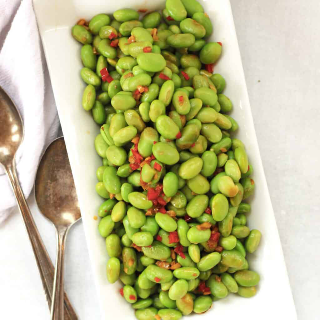 Cooked edamame beans on a white plate next to two spoons.