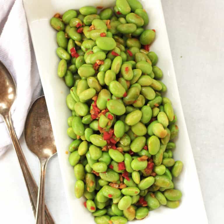 Cooked edamame beans on a white plate next to two spoons.