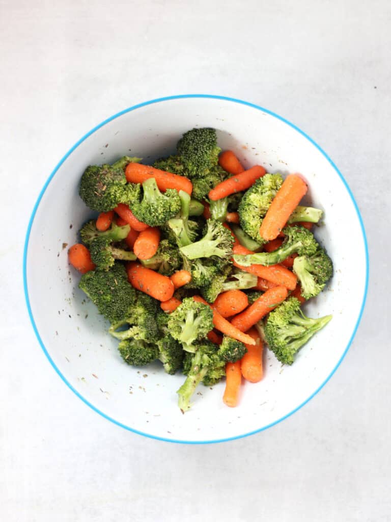 Carrots and broccoli tossed with oil and seasoning in a mixing bowl.