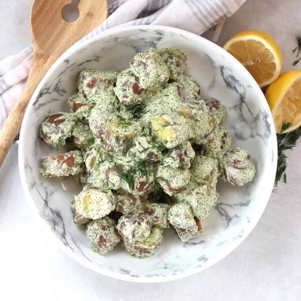 A bowl of potato salad next to a fresh lemon, herbs and salad server.