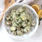 A bowl of potato salad next to a fresh lemon, herbs and salad server.