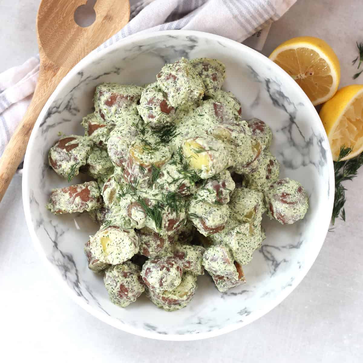 A bowl of potato salad next to a fresh lemon, herbs and salad server.