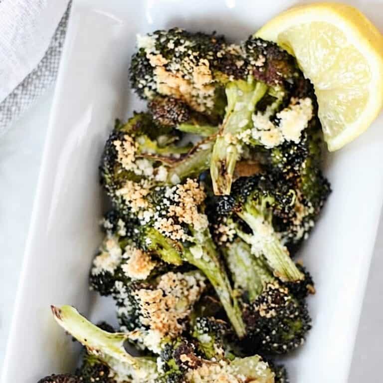 Oven roasted crispy broccoli served on a white plate with parmesan topping and a slice of lemon.