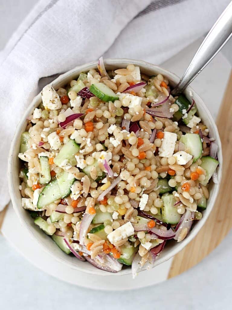 Pearl couscous with feta, cucumber and red onion in a serving bowl with a spoon.